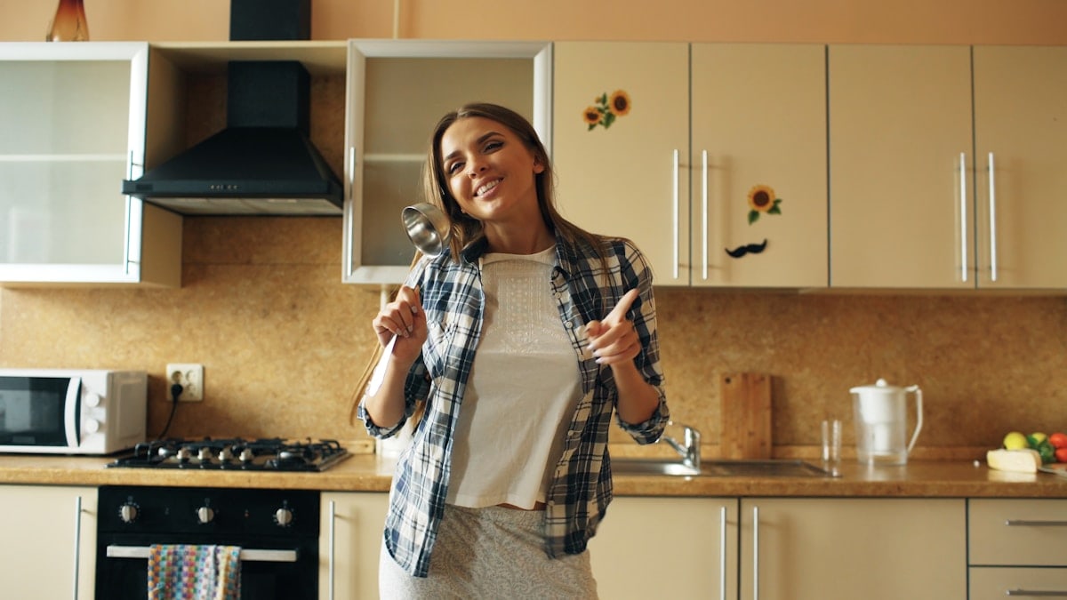 Cheerful young funny woman dancing and singing with ladle while having leisure time in the kitchen at home in the morining